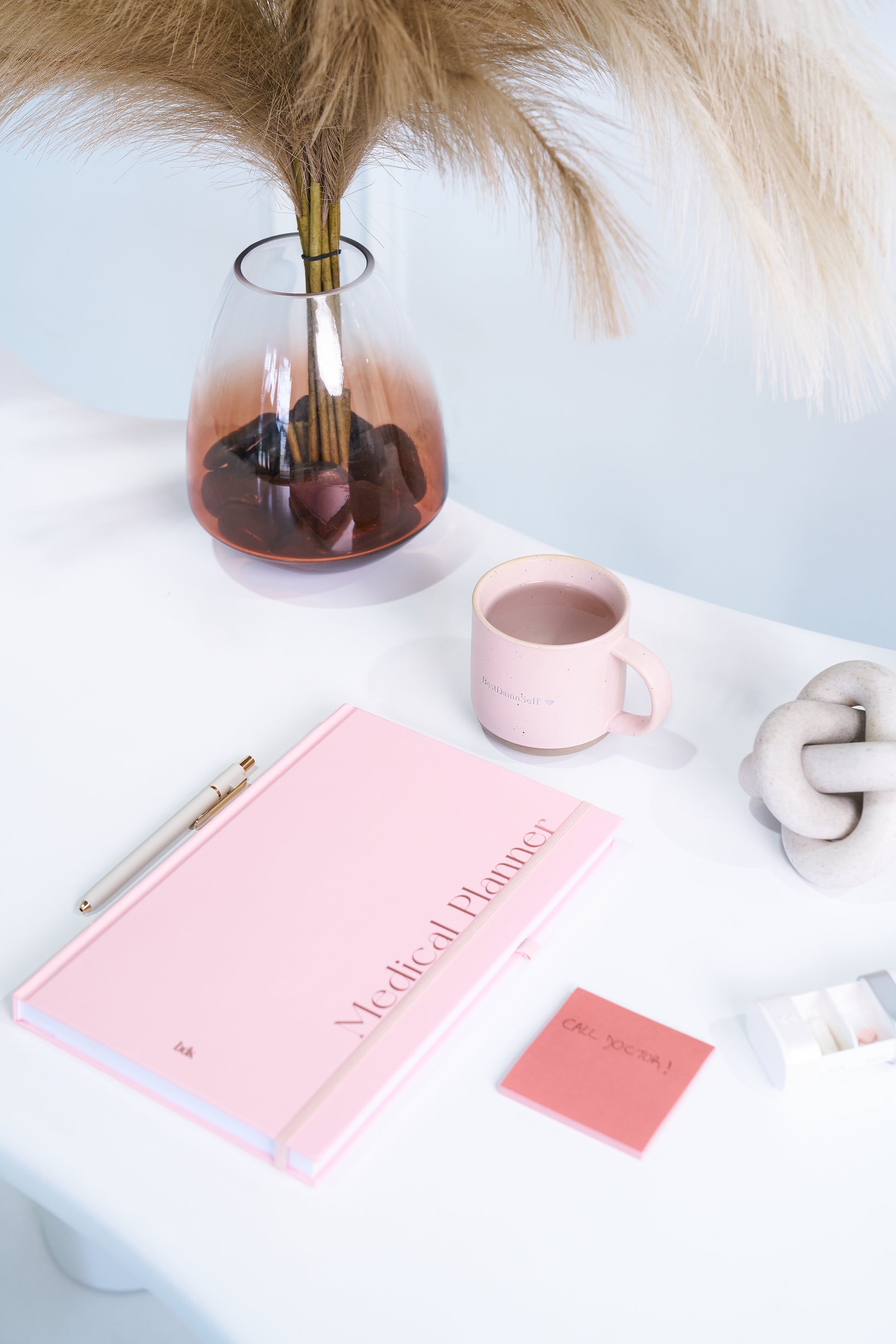 A stylish workspace with a pink medical planner, matching mug, and note card on a white desk