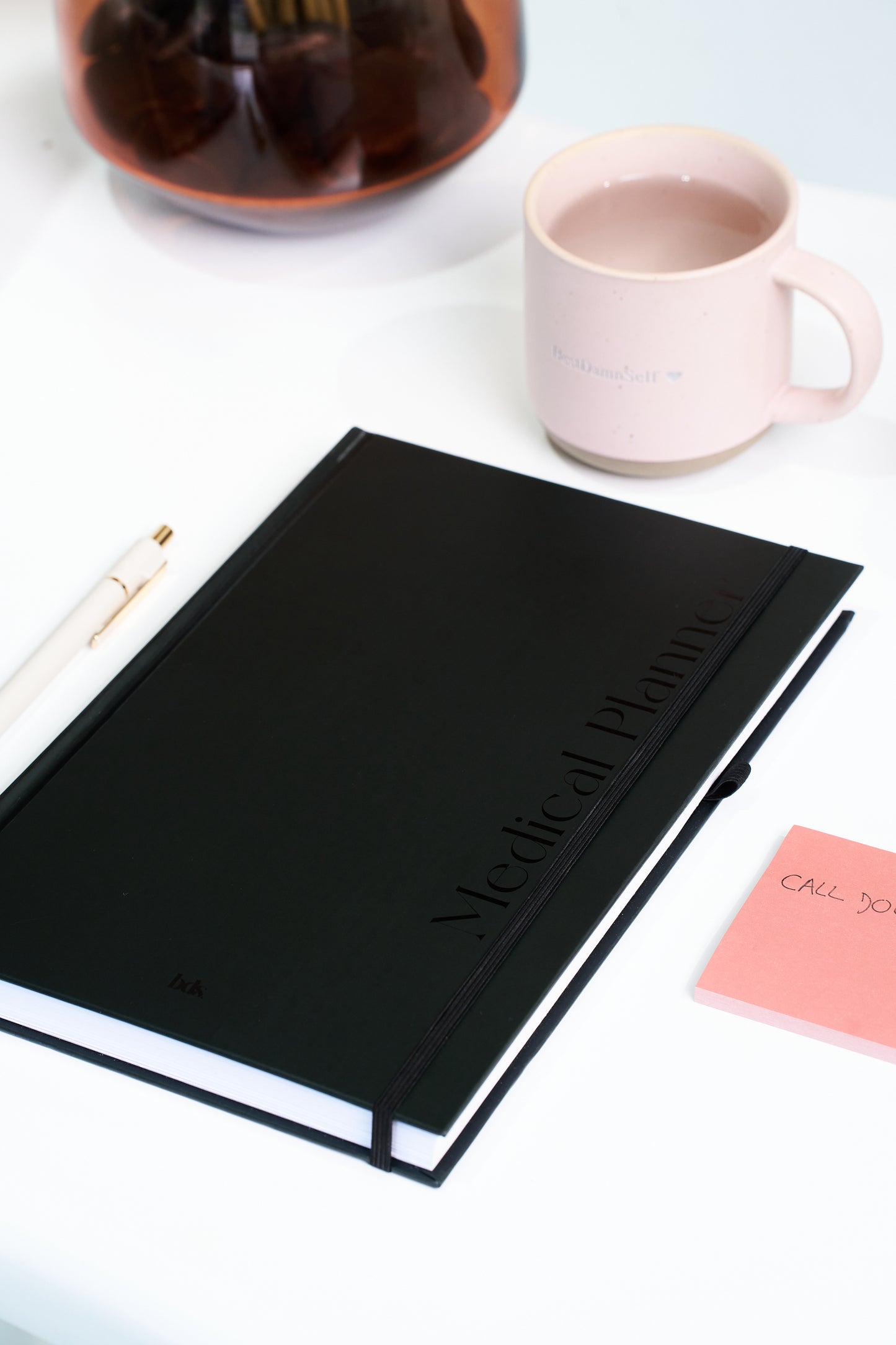 A black medical planner, white pen, pink sticky note, and pink mug sit on a white table