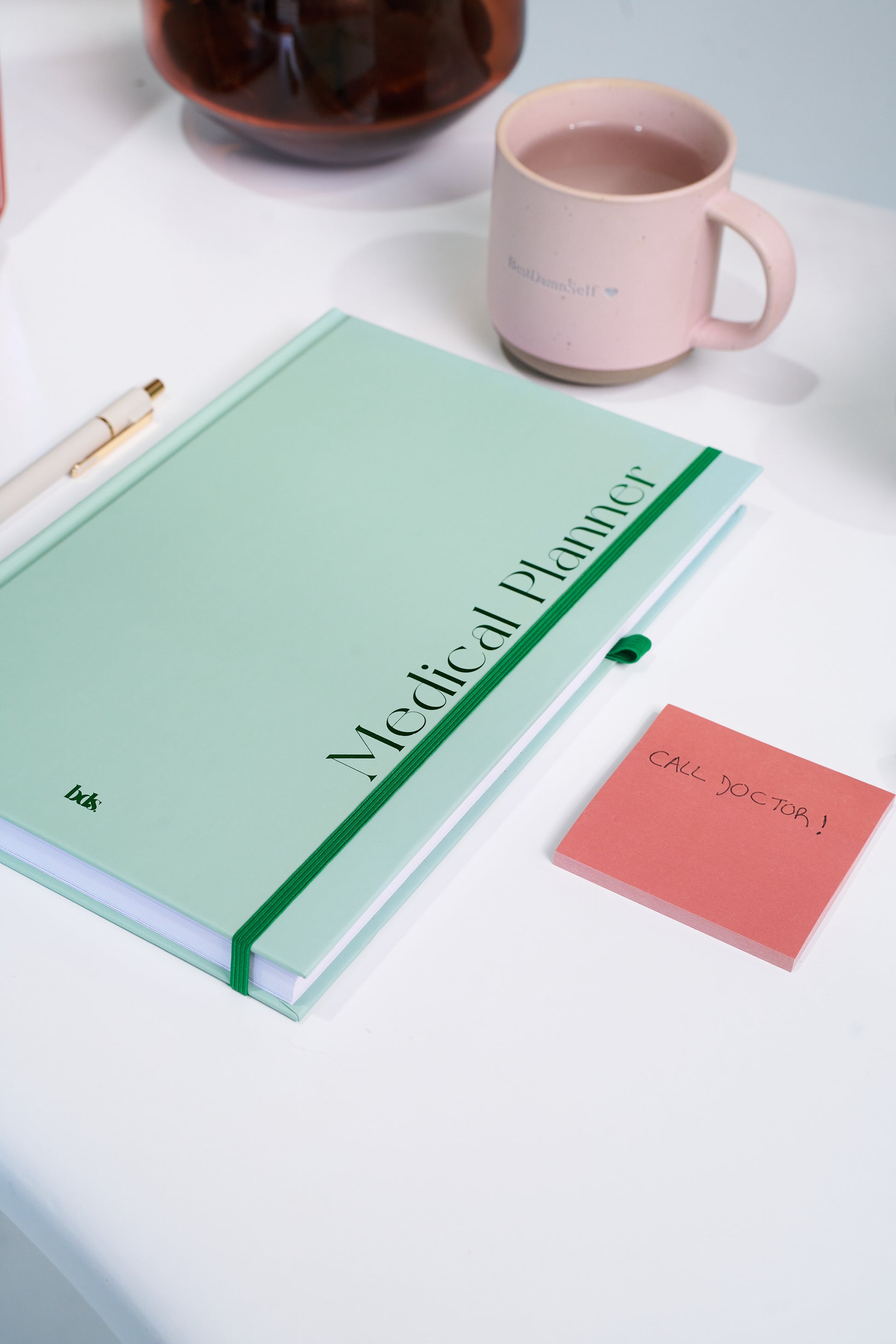 A mint green medical planner with a pen rests on a white table. Beside it, a pink mug and a note reading "CALL DOCTOR" are visible