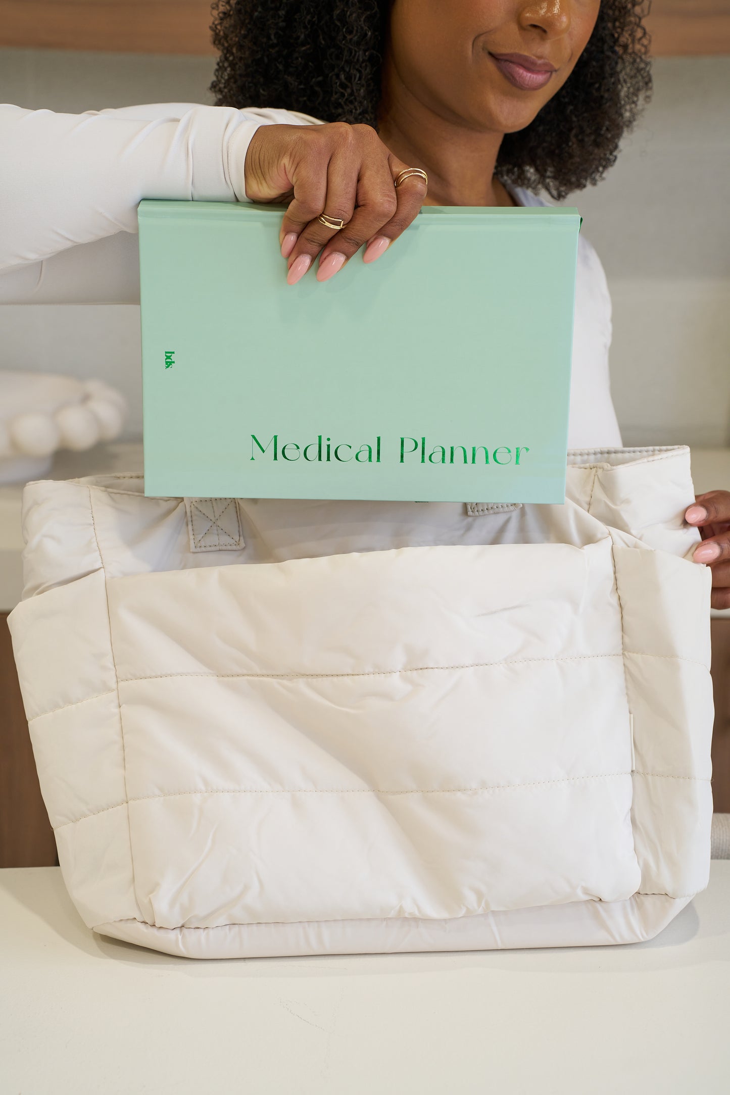 A person is placing a light green "Medical Planner" into a large, quilted white bag on a table.