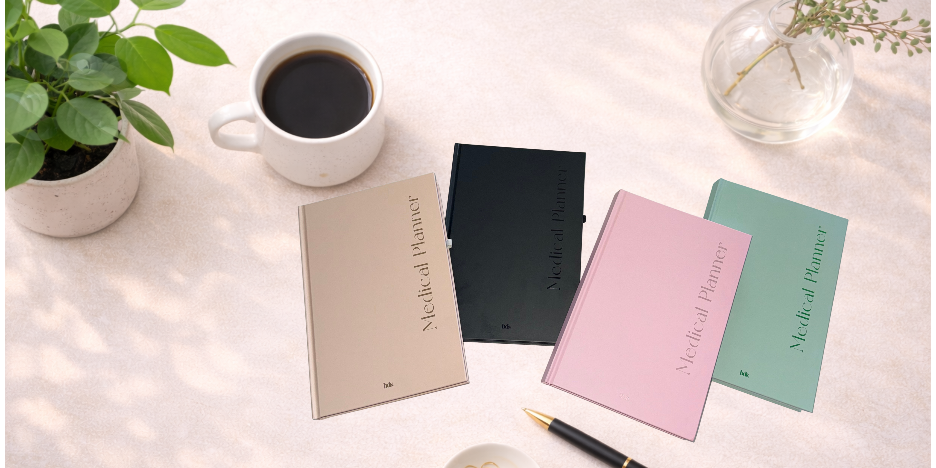 A table with four medical planners in beige, black, pink, and mint green covers. A coffee cup, a plant, and a pen